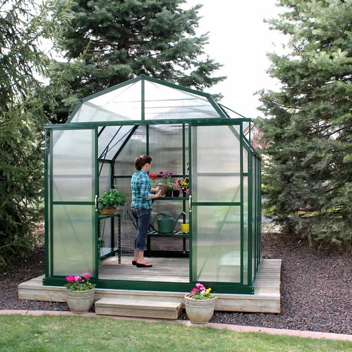 Grandio Elite Greenhouse on wooded platform next to pine trees with plants getting watered inside 